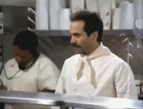 A Man In A White Shirt And Tie Is Standing In A Kitchen Next To A Woman Talking On A Phone . GIF