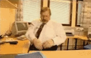A Man In A White Shirt And Tie Sits At A Desk GIF