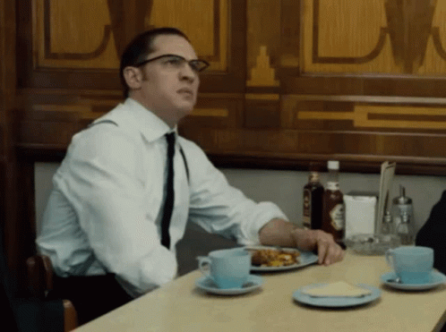 A Man In A White Shirt And Tie Sits At A Table With Plates Of Food GIF