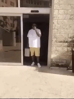 A Man In A White Shirt And Yellow Shorts Is Standing In A Doorway . GIF