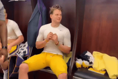 A Man In A White Shirt And Yellow Shorts Sits In A Locker Room . GIF