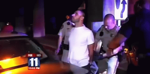 A Man In A White Shirt Is Being Escorted By Police Officers In Front Of A Fox 11 News Car GIF