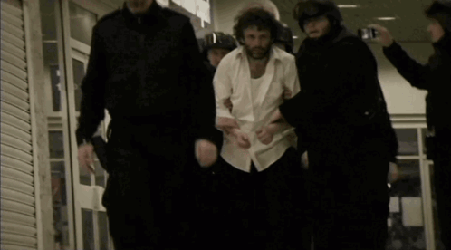 A Man In A White Shirt Is Being Led By A Group Of Police Officers GIF
