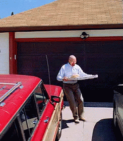 A Man In A White Shirt Is Carrying A Pizza In Front Of A Garage Door GIF