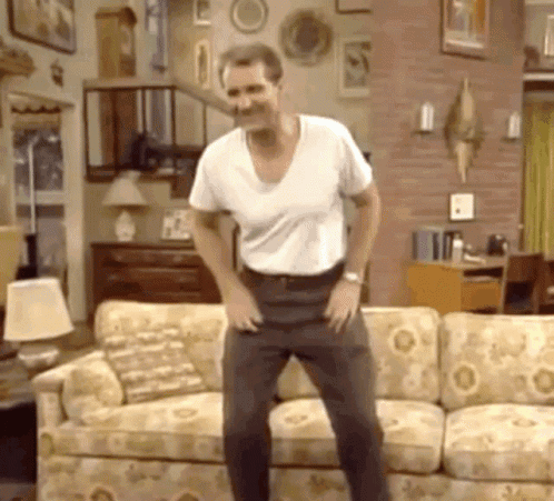 A Man In A White Shirt Is Dancing In Front Of A Couch In A Living Room GIF