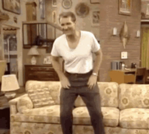 A Man In A White Shirt Is Dancing In Front Of A Couch . GIF