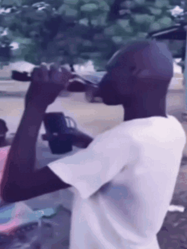 A Man In A White Shirt Is Drinking From A Bottle Outside GIF