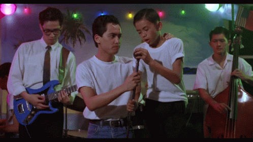 A Man In A White Shirt Is Holding A Microphone While Another Man Plays A Guitar GIF