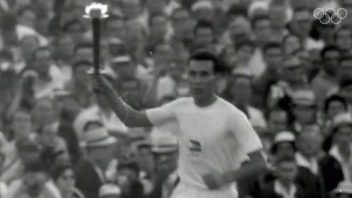 a man in a white shirt is holding a torch in front of a crowd with the olympic rings in the background