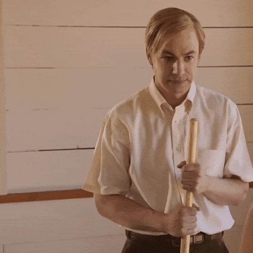 A Man In A White Shirt Is Holding A Wooden Stick In His Right Hand GIF