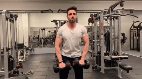 A Man In A White Shirt Is Holding Dumbbells In A Gym . GIF