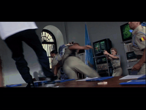 A Man In A White Shirt Is Kicking Another Man In A Room With Other People GIF