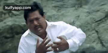 A Man In A White Shirt Is Making A Funny Face While Sitting On A Rock . GIF