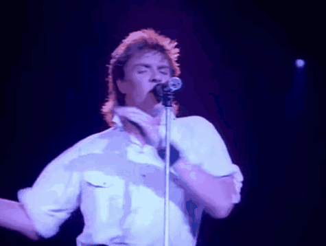 a man in a white shirt is singing into a microphone and making a peace sign