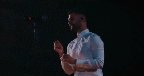 A Man In A White Shirt Is Singing Into A Microphone In A Dark Room GIF
