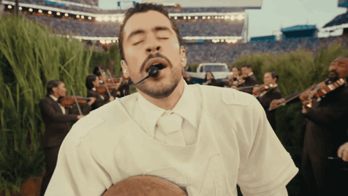 A Man In A White Shirt Is Singing Into A Microphone In Front Of A Band GIF