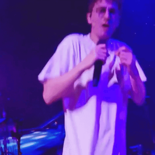 A Man In A White Shirt Is Singing Into A Microphone On Stage GIF