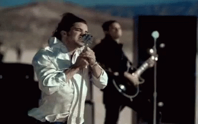 A Man In A White Shirt Is Singing Into A Microphone While Another Man Plays A Guitar . GIF