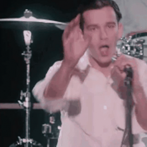 A Man In A White Shirt Is Singing Into A Microphone While Standing In Front Of A Drum Set . GIF