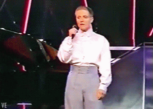 A Man In A White Shirt Is Singing Into A Microphone While Standing On A Stage GIF