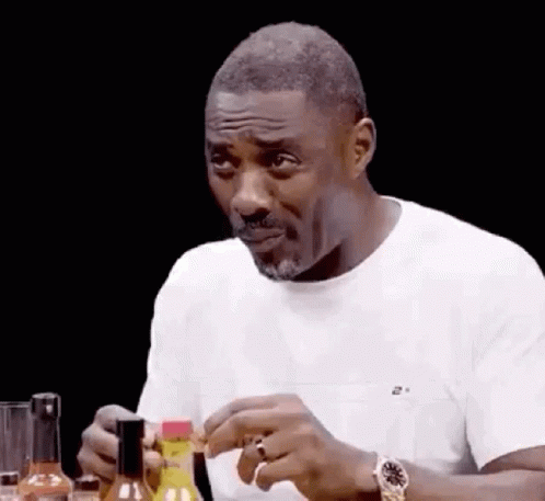 A Man In A White Shirt Is Sitting At A Table With A Bottle Of Hot Sauce In His Hand . GIF