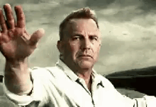 A Man In A White Shirt Is Sitting In A Car Waving His Hand . GIF