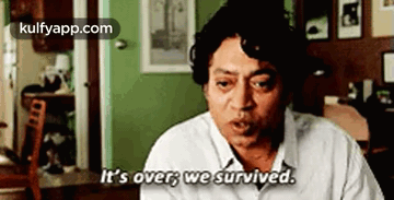 A Man In A White Shirt Is Sitting In A Living Room And Saying `` It 'S Over We Survived '' . GIF