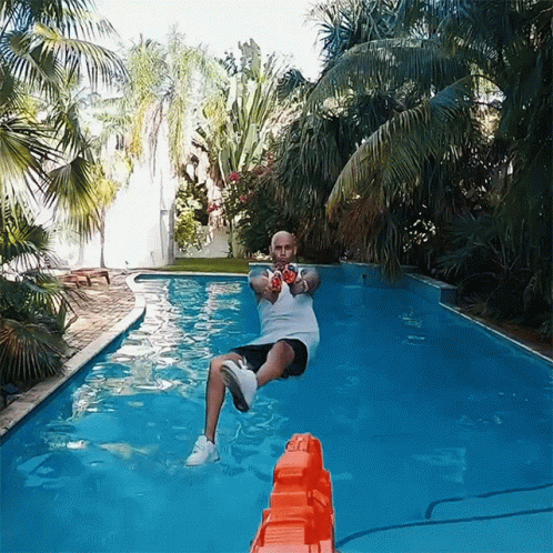 A Man In A White Shirt Is Sitting In A Swimming Pool Holding A Nerf Gun GIF