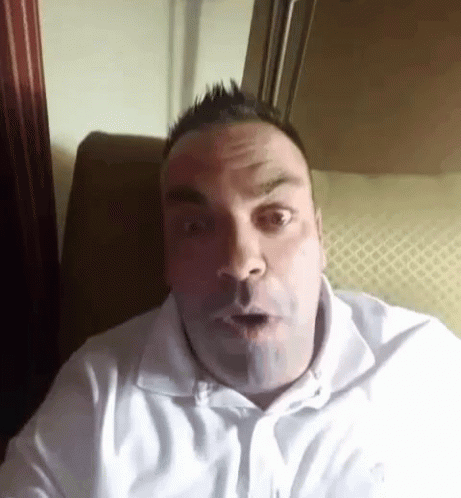a man in a white shirt is sitting on a couch and making a surprised face .
