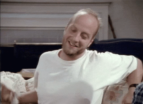 A Man In A White Shirt Is Sitting On A Couch Smiling GIF