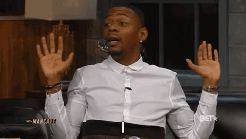 A Man In A White Shirt Is Sitting On A Couch With His Hands In The Air GIF