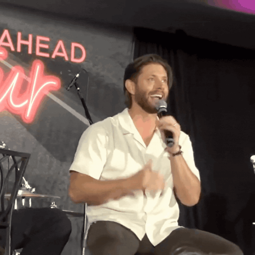 A Man In A White Shirt Is Speaking Into A Microphone In Front Of A Sign That Says Ahead GIF