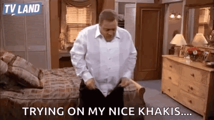 A Man In A White Shirt Is Standing In A Bedroom . GIF