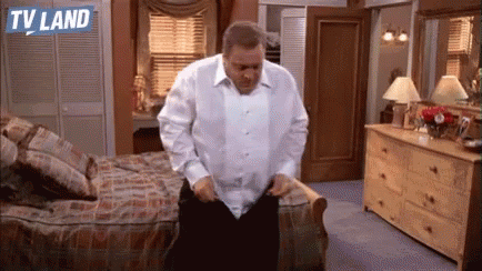 A Man In A White Shirt Is Standing In A Bedroom With A Dresser And A Bed . GIF