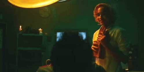 A Man In A White Shirt Is Standing In A Dark Room Holding A Glass Of Wine . GIF