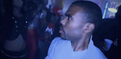A Man In A White Shirt Is Standing In A Dark Room With A Crowd Behind Him . GIF