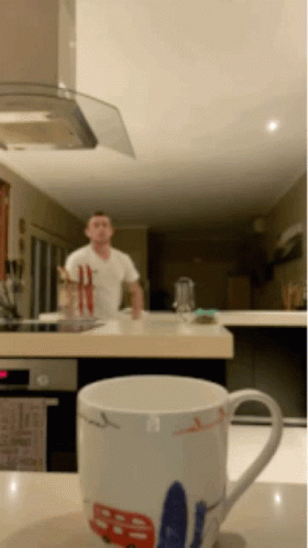 A Man In A White Shirt Is Standing In A Kitchen Behind A Mug That Says ' Sd ' On It GIF