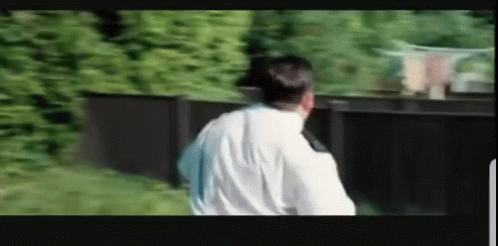 A Man In A White Shirt Is Standing In Front Of A Black Fence . GIF