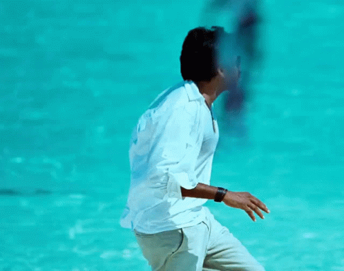 A Man In A White Shirt Is Standing In Front Of A Blue Water GIF
