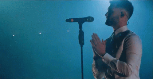 A Man In A White Shirt Is Standing In Front Of A Microphone Covering His Face . GIF