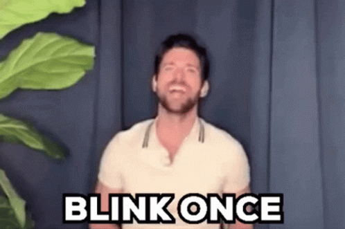 A Man In A White Shirt Is Standing In Front Of A Plant And Saying Blink Once . GIF