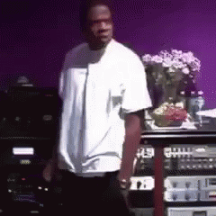 A Man In A White Shirt Is Standing In Front Of A Purple Wall With A Vase Of Flowers In The Background . GIF