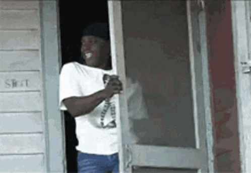 A Man In A White Shirt Is Standing In Front Of A Screen Door Holding A Drink . GIF