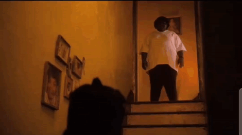 A Man In A White Shirt Is Standing On The Stairs In A Dark Room . GIF