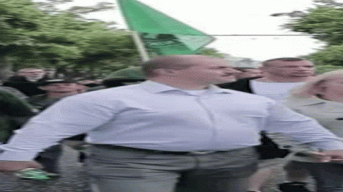 A Man In A White Shirt Is Walking Down A Street With A Green Flag In His Hand . GIF