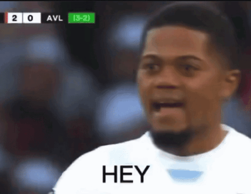 A Man In A White Shirt Says Hey In Front Of A Scoreboard GIF