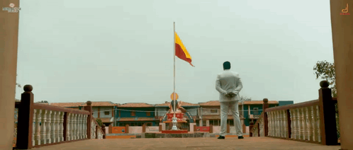 A Man In A White Shirt Stands In Front Of A Building With A Flag Flying In The Background GIF