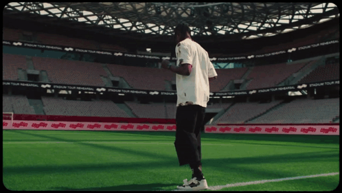 A Man In A White Shirt Stands On A Soccer Field In Front Of A Stadium That Says Stadium Tour GIF