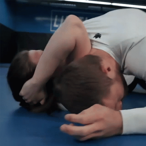A Man In A White Shirt With An Arrow On It Is Wrestling Another Man On A Blue Mat GIF