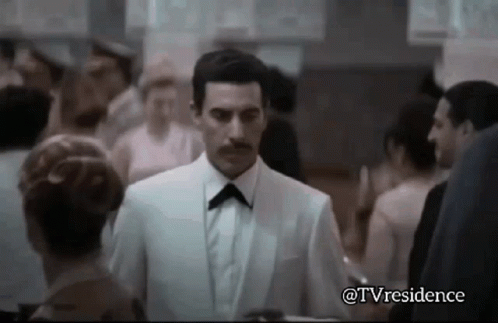 A Man In A White Suit And Black Bow Tie Is Walking Through A Crowd Of People . GIF